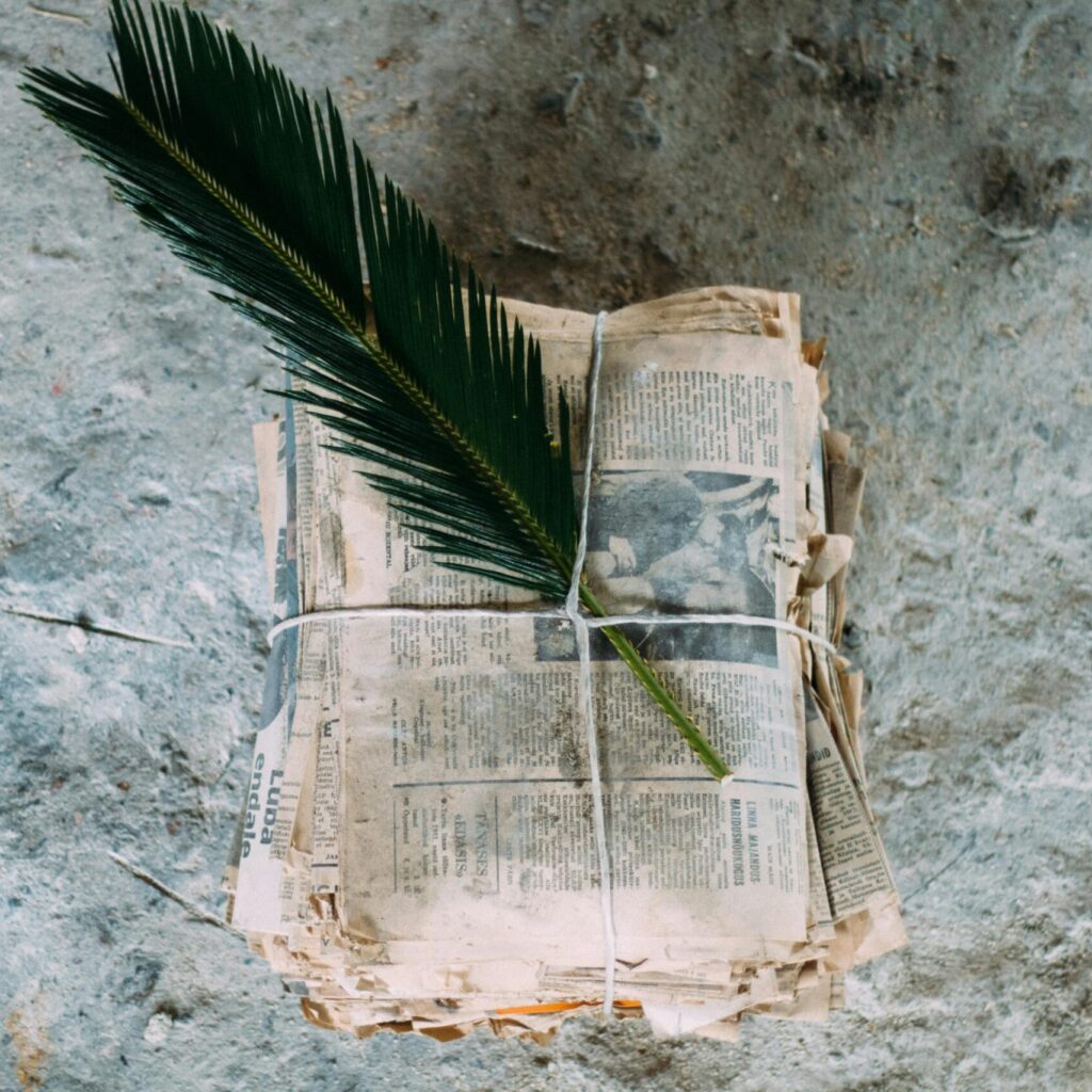 Overhead view of a stack of old newspapers tied with string, topped with a lush green palm leaf on a concrete floor.